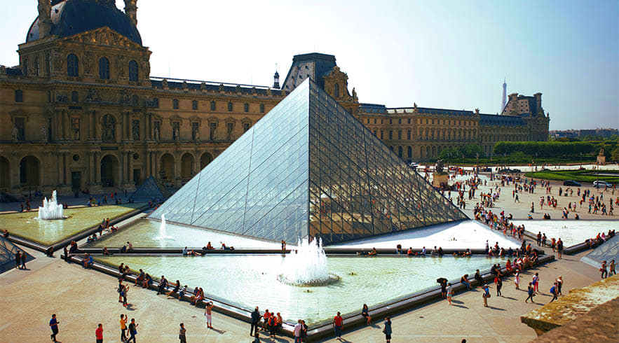 Glass pyramid entrance to the Louvre Museum in Paris with visitors in the courtyard and classical palace buildings in backgro